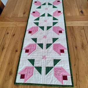 May include: A rectangular quilted table runner with a repeating pattern of pink tulip shapes and green leaves on a white background. The runner has a green border and is placed on a wooden table.