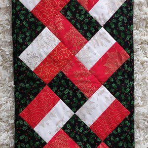 May include: A quilted table runner featuring a geometric pattern of red, white, and black fabric squares. The red and white squares are decorated with gold-toned botanical and swirl prints, while the black squares feature a green holly leaf pattern.