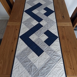 May include: A white and blue quilted table runner with a geometric pattern. The runner features a series of overlapping squares in a navy blue colour.