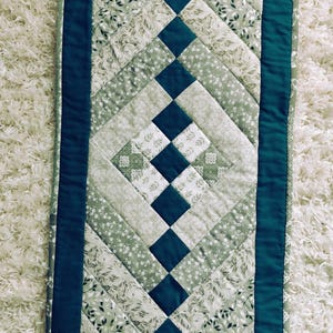 May include: A quilted table runner featuring a geometric pattern of dark teal solid fabric and light green floral print fabric. The design consists of a central column of dark teal squares flanked by diagonal strips of various green floral patterns.