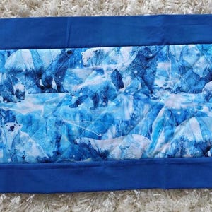 May include: A rectangular table runner featuring a central panel with a repeating pattern of white polar bears on a blue icy background, framed by a solid royal blue fabric border with pointed ends on both sides of the textile piece.