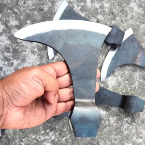 May include: A hand holds a black metal axe with a curved blade. The axe has a sharp edge and a rounded handle. The axe is on a gray concrete surface.