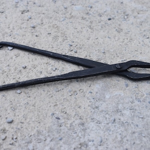 Może przedstawiać: Szczypce kowalskie, narzędzie ręczne z długimi uchwytami i chwytającą głowicą, leżą na teksturowanej, szarej powierzchni. Szczypce wykonane są z ciemnego metalu, z zakrzywionymi pętlami na końcach uchwytów.