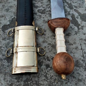 May include: A Roman Gladius sword with a wooden handle and a brass scabbard. The sword has a double-edged blade and a leather grip.