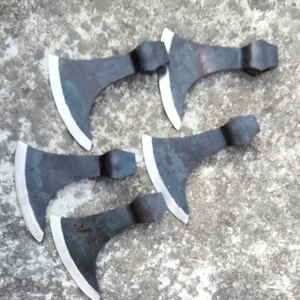 May include: Five hand-forged axes with black metal heads and polished silver blades. The axes are arranged in a semi-circle on a gray concrete surface.