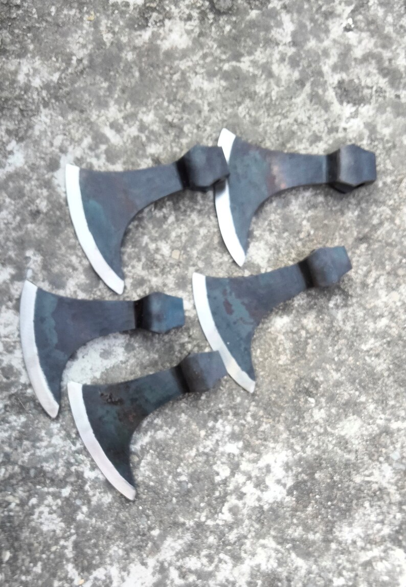 May include: Five hand-forged axes with black metal heads and polished silver blades. The axes are arranged in a semi-circle on a gray concrete surface.