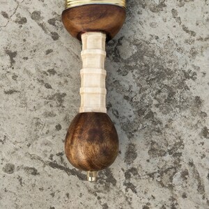 May include: Close-up of the pommel of a sword with a wooden handle and a brass hilt. The handle is made of light brown wood and has a white bone-like grip. The pommel is rounded and also made of wood.