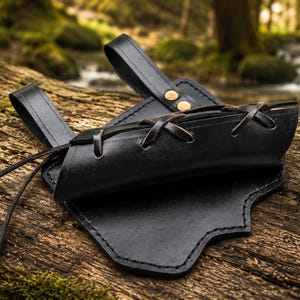 May include: A black leather sheath featuring a lace-up design with thin leather cords and two belt loops secured by two brass-colored metal rivets. The sheath has a curved, contoured shape with visible stitching along the edges of the leather.