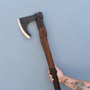 以下が含まれることがあります： 手は、黒い刃と革の巻き付けが施された茶色の柄の斧を持っています。斧の柄には彫刻が施されています。