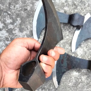 May include: A hand holds a black metal axe with a curved blade and a square handle. The axe is made of forged steel and has a sharp edge. The axe is a traditional weapon and tool.