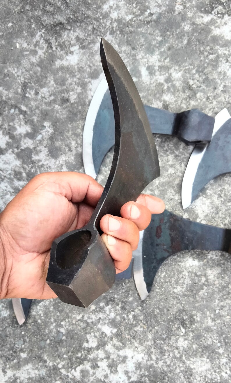 May include: A hand holds a black metal axe with a curved blade and a square handle. The axe is made of forged steel and has a sharp edge. The axe is a traditional weapon and tool.