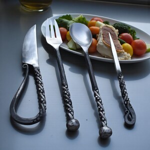 May include: Hand-forged metal cutlery set including a knife, fork, spoon, and butter knife. The handles have a twisted rope design, and the knife has a loop at the end. The set is displayed near a plate of food.