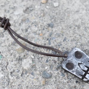 May include: A silver-colored pendant necklace with a skull design. The pendant is rectangular with two circular eye holes and a jagged mouth. The necklace has a brown leather cord.
