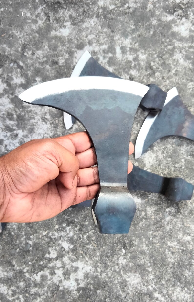 May include: A hand holds a black metal axe with a curved blade. The axe has a sharp edge and a rounded handle. The axe is on a gray concrete surface.