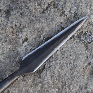 Könnte beinhalten: Nahaufnahme eines Speers mit einer scharfen, dreieckigen Klinge. Die Metallklinge hat eine dunkle, matte Oberfläche mit einer helleren Kante und liegt auf einer strukturierten, grauen Steinoberfläche. Der Speer ist eine Waffe.