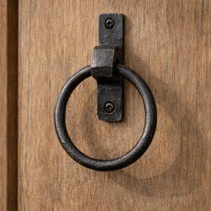 May include: A black, wrought iron ring pull door handle mounted on a wooden surface. The handle is circular and attached to a rectangular plate with two screws. The wood has a natural grain.