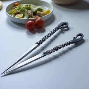 Op de afbeelding: Twee handgemaakte metalen spiesen met gedraaide handvatten en puntige uiteinden. De spiesen hebben een donkere, getextureerde afwerking en een decoratieve lus aan het einde van het handvat. Een kom salade en twee rode tomaten staan op de achtergrond.