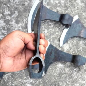 May include: Close-up of the handle of a black axe with a rounded hole in the center. The axe is held in a hand, and there are other axes in the background.