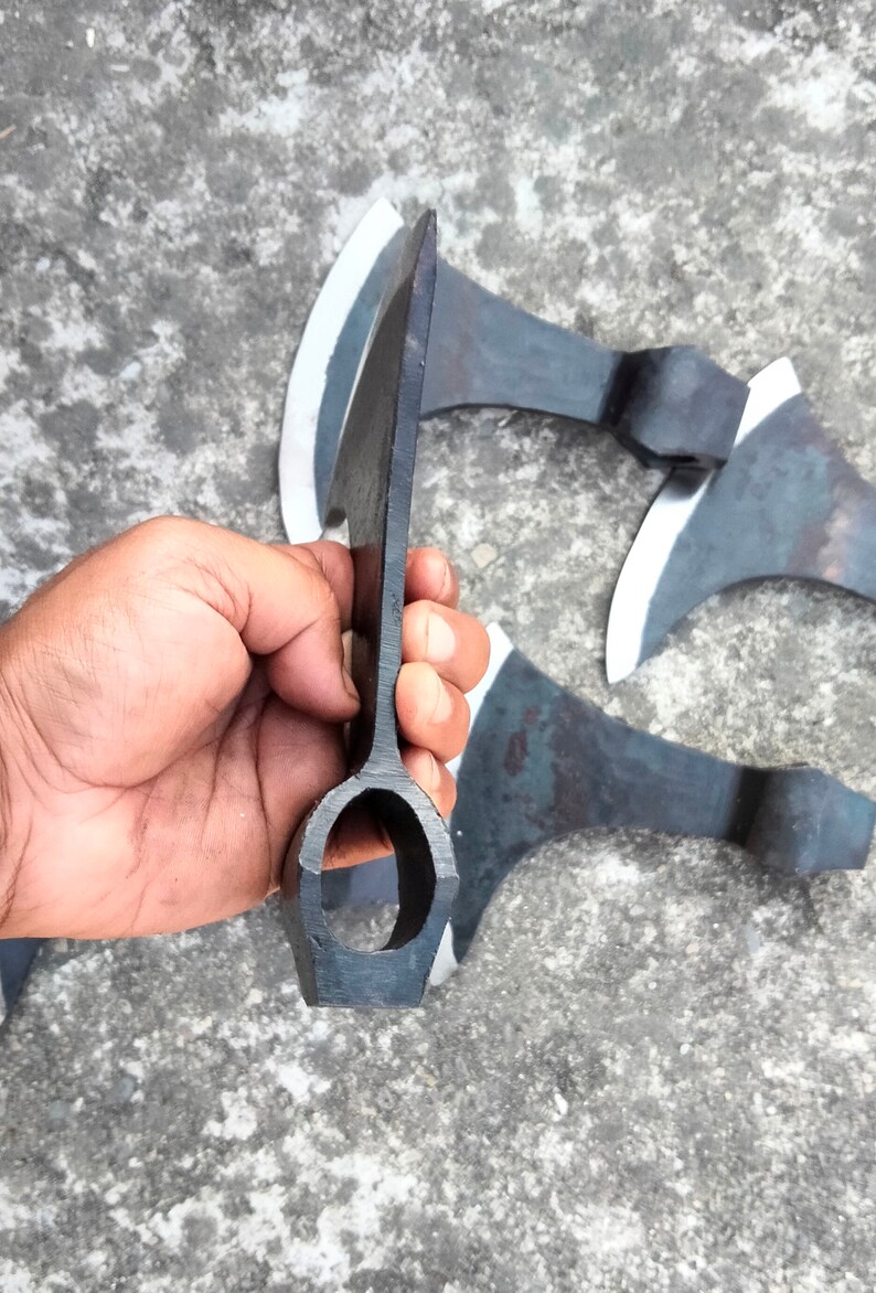 May include: Close-up of the handle of a black axe with a rounded hole in the center. The axe is held in a hand, and there are other axes in the background.