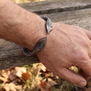 Puede incluir: Una pulsera de metal artesanal, abierta, con un acabado gris oscuro texturizado. La pulsera presenta un diseño curvo estilizado con elementos decorativos. La pulsera se muestra en la muñeca de una persona.