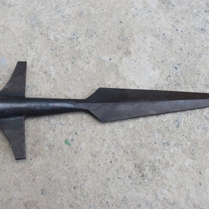 May include: A black metal spearhead with a wide base and a sharp point. The spearhead is lying on a gray concrete surface.