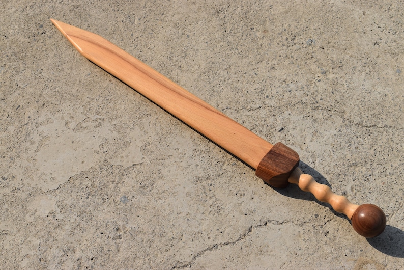K&ouml;nnte beinhalten: Ein Holzschwert mit einem braunen Griff und einem abgerundeten Knauf. Das Schwert ist aus hellem Holz gefertigt und hat eine scharfe Spitze.