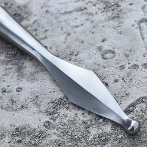 Puede incluir: Una punta de lanza plateada pulida, con una hoja en forma de diamante y una base redondeada. La punta de lanza es de metal y está colocada sobre una superficie de hormigón gris texturizado.