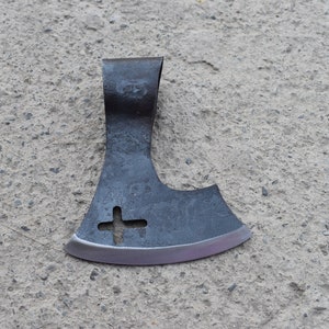 May include: A black axe head with a cross-shaped hole in the blade. The axe head is lying on a gray concrete surface.