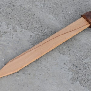 K&ouml;nnte beinhalten: Ein h&ouml;lzernes Spielzeugschwert mit einem braunen Holzgriff und einer hellbraunen Holzklinge.