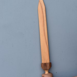 K&ouml;nnte beinhalten: Ein Holzschwert mit einem braunen Griff und einer hellbraunen Klinge. Das Schwert wird von einer Person mit einem Tattoo am Arm gehalten.