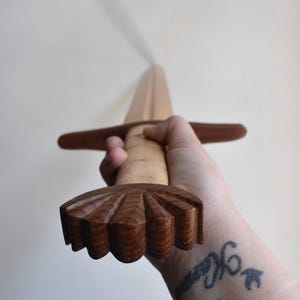 Puede incluir: Una espada de madera con una hoja de color claro y una empuñadura y guarda de color marrón oscuro. El mango tiene un diseño decorativo tallado. La espada se sostiene en una mano.