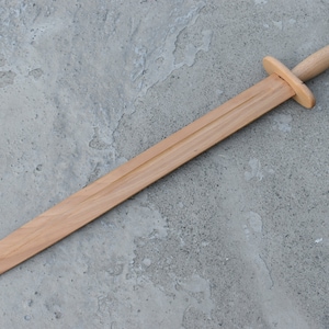 Puede incluir: Una espada de juguete de madera con una punta redondeada y un diseño simple. La espada está hecha de madera marrón claro y mide aproximadamente 60 cm de largo.