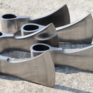 May include: A collection of five unhandled axe heads made of silver-colored metal. The axe heads are arranged in a slightly overlapping pattern, with the blades facing the same direction. The axe heads are on a concrete surface.