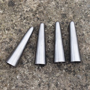 Puede incluir: Cuatro conos de metal plateado, cada uno con una punta puntiaguda y una superficie lisa y pulida. Los conos están dispuestos en una fila sobre una superficie gris texturizada.