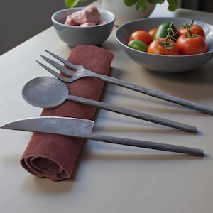 May include: Hand-forged metal flatware set including a fork, spoon, and knife. The utensils are placed on a folded burgundy napkin. Bowls of food, including tomatoes and meat, are in the background.