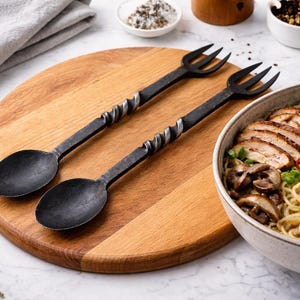 May include: Two black metal serving utensils, a spoon and a fork, rest on a wooden cutting board. A bowl of ramen with sliced chicken, mushrooms, and eggs sits nearby. Rosemary sprigs and spices are also visible.