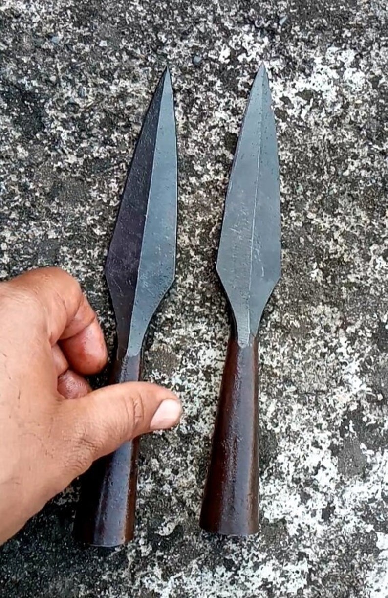 May include: Two dark grey metal spearheads with brown wooden handles. The spearheads are pointed and have a slightly curved shape.