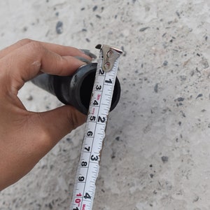May include: A white measuring tape with black markings is used to measure the diameter of a black pipe. The tape reads 2.5 cm.