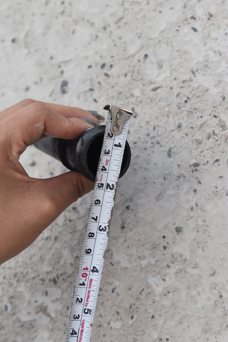 May include: A white measuring tape with black markings is used to measure the diameter of a black pipe. The tape reads 2.5 cm.