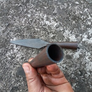 May include: A close-up of a hand holding a brown metal spear with a sharp point. The spear is lying on a grey concrete surface.