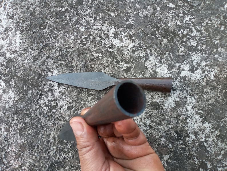 May include: A close-up of a hand holding a brown metal spear with a sharp point. The spear is lying on a grey concrete surface.