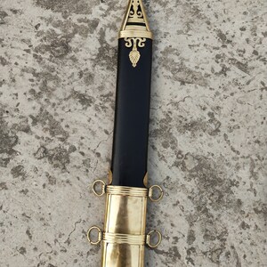 May include: A black scabbard with gold accents and a decorative top. The scabbard has two rings on each side.
