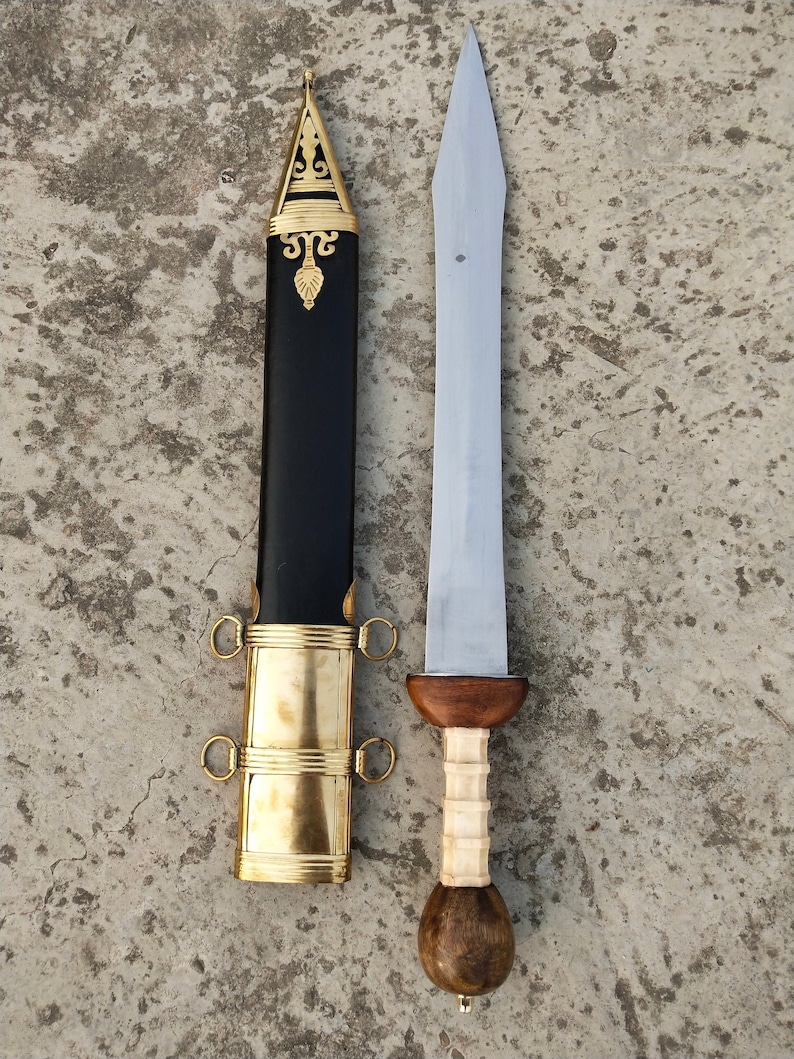 May include: A Roman gladius sword with a wooden handle and a metal blade. The sword is in a scabbard with gold accents.