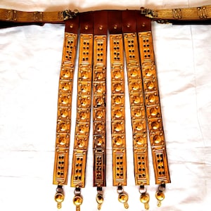 May include: A gold-colored leather belt with multiple rows of decorative metal plates. The belt has a buckle and a series of hanging tassels.