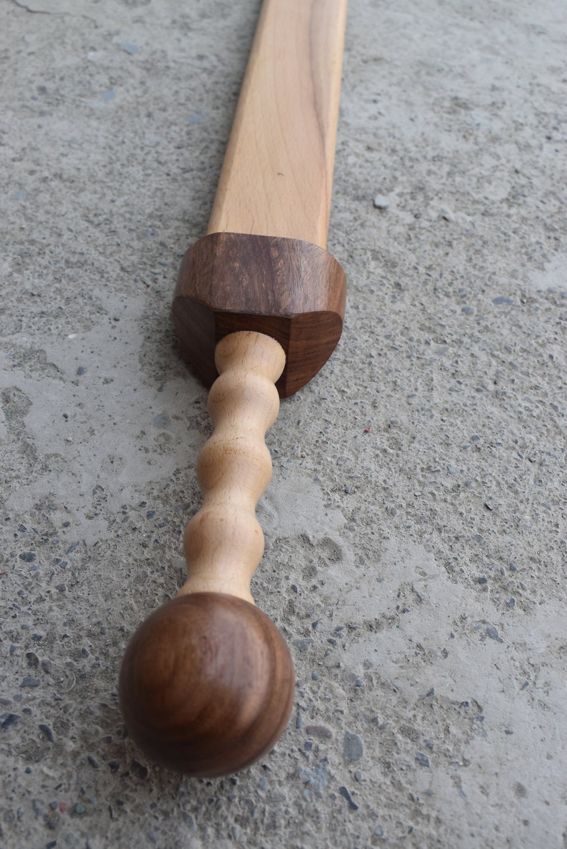 K&ouml;nnte beinhalten: Ein Holzschwert mit einem braunen Griff und einer hellbraunen Klinge. Der Griff hat einen runden Knopf am Ende und ein gedrehtes Design in der Mitte.