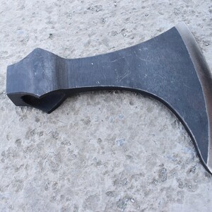 May include: A black metal axe with a curved blade and a handle with a hole in the top. The axe is lying on a gray concrete surface.
