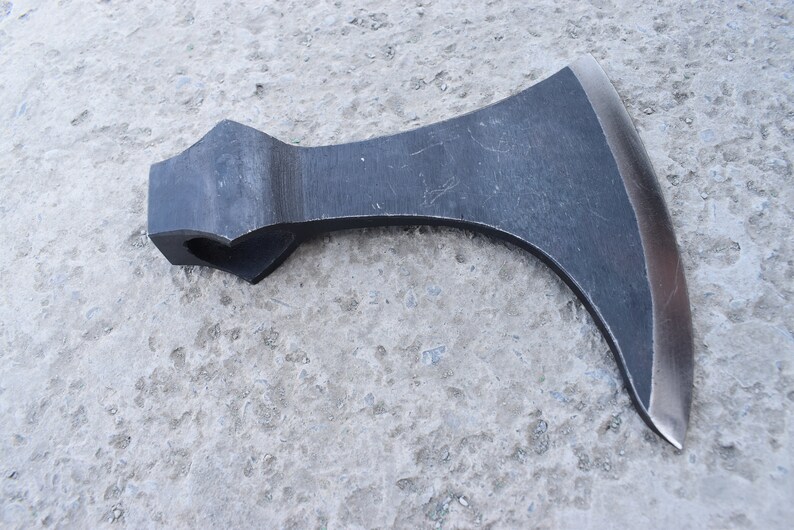 May include: A black metal axe with a curved blade and a handle with a hole in the top. The axe is lying on a gray concrete surface.