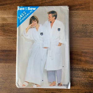 May include: A vintage sewing pattern titled "See & Sew 5452" featuring a couple wearing white robes. The pattern includes sizes A(XS-S-M-L-XL). The robes have a shawl collar, a belt, and a small emblem on the chest.