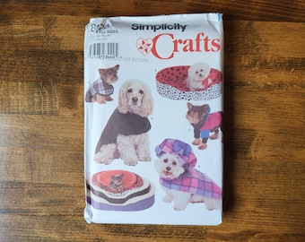 May include: Simplicity Crafts sewing pattern for dog clothes and a dog bed. The pattern includes instructions for making a plaid dog coat, a black dog coat, a red and black dog coat, and a dog bed with a black and white polka dot pattern. The pattern is for all sizes.