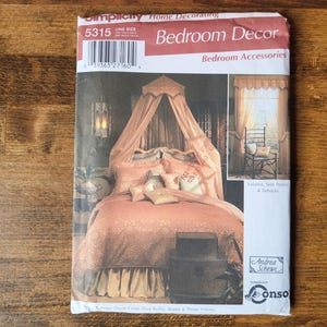 May include: A Simplicity 5315 sewing pattern envelope for bedroom decor, featuring a photograph of a bed with a peach-coloured canopy, matching bedding, and pillows. The package includes text for valance, side panels, and tiebacks, with a barcode.